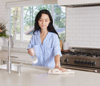 A woman standing at a kitchen counter, using a Glass Branch Basics bottle and a cloth towel to clean the counter's surface. - Thumbnail View 6
