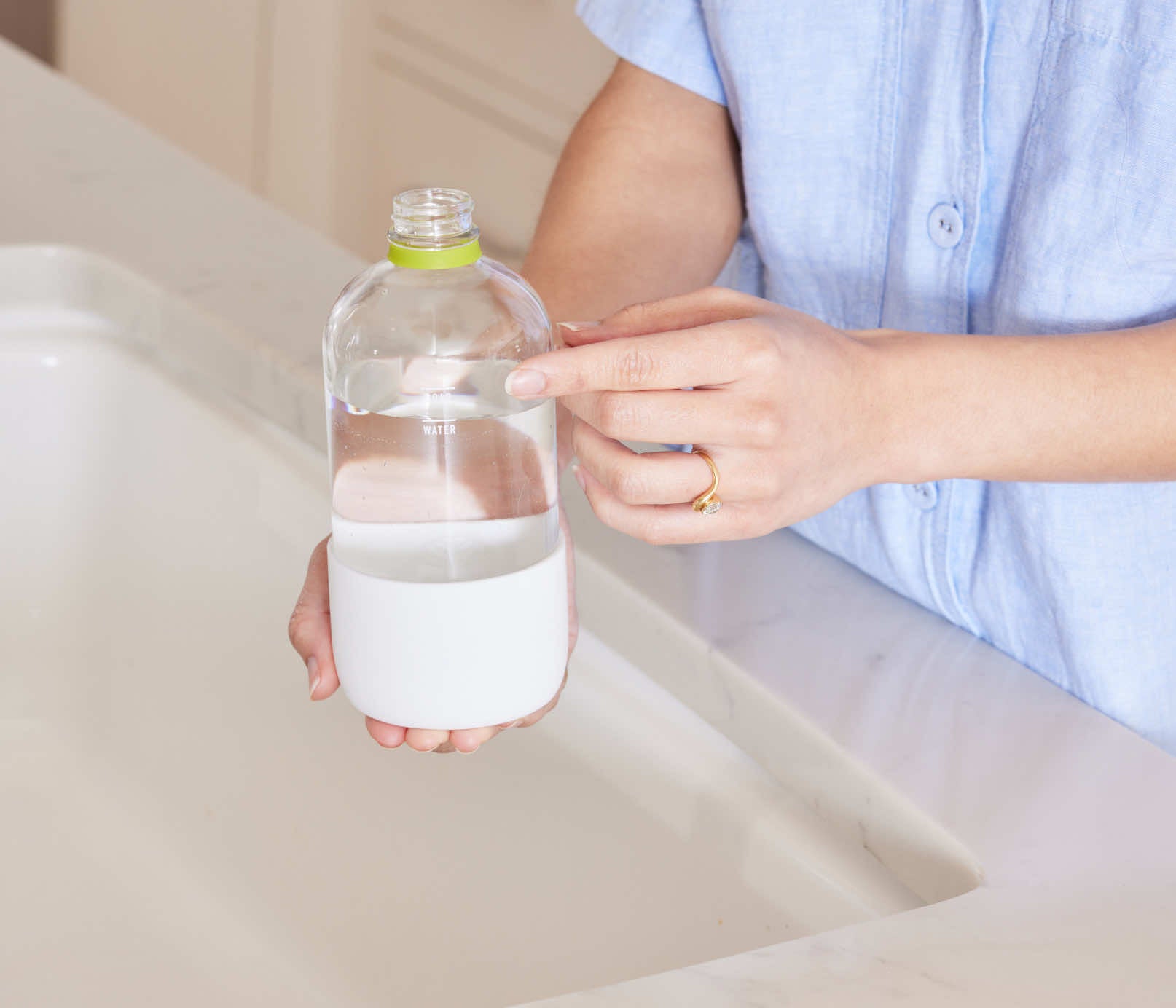 A woman standing at a kitchen faucet, pointing at the fill-lines on a refillable Glass Branch Basics bottle; there are separate fill-lines for water and Branch Basics Concentrate, respectively. - View 4
