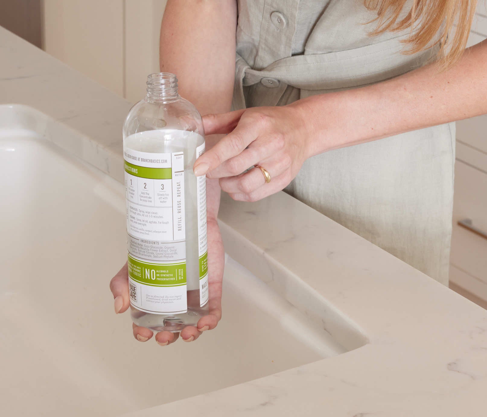 A woman standing at a kitchen faucet, pointing at the fill-lines on a refillable Plastic Branch Basics bottle; there are separate fill-lines for water and Branch Basics Concentrate, respectively. - View 3
