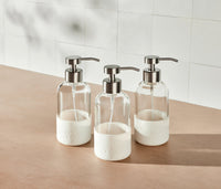 A trio of refillable Glass Foaming Wash Bottles with Brushed-Chrome pumps, displayed on a neutral surface and white tiled background. - Thumbnail View 2
