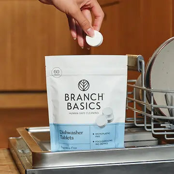 Branch Basics Dishwasher Tablets packaging with a hand holding a tablet above it, set against a kitchen background.