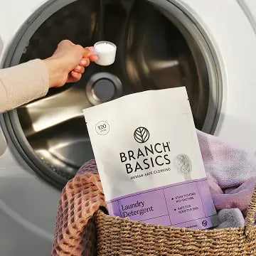 Person holding Branch Basics Laundry Detergent next to a washing machine with a basket of clothes.