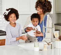 A single refillable Glass Foaming Wash Bottle with a Brushed-Chrome pump being used by a mom and two children to wash their hands in a kitchen sink. - Thumbnail View 3
