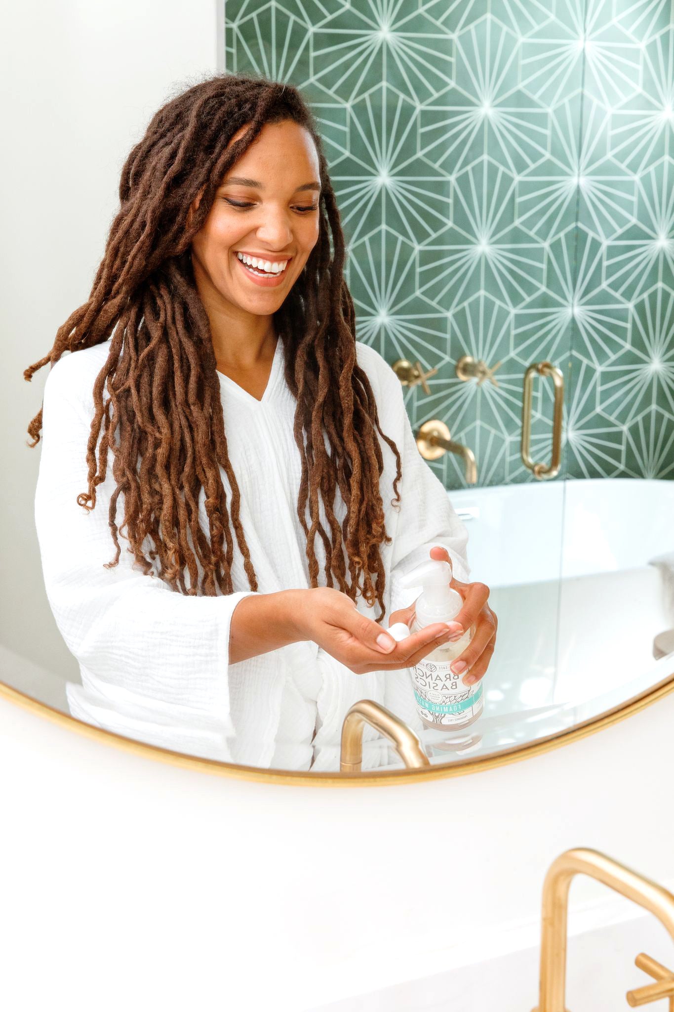 A person shown in a mirror standing at a sink, using a refillable Plastic Foaming Wash Bottle to dispense soap into their hands. - View 3
